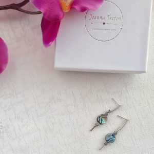kolczyki z muszlą abalone leżą na obrusie, nad nimi opakowanie z napisem "Joanna Trefon, handmade jewellery" i kwiat